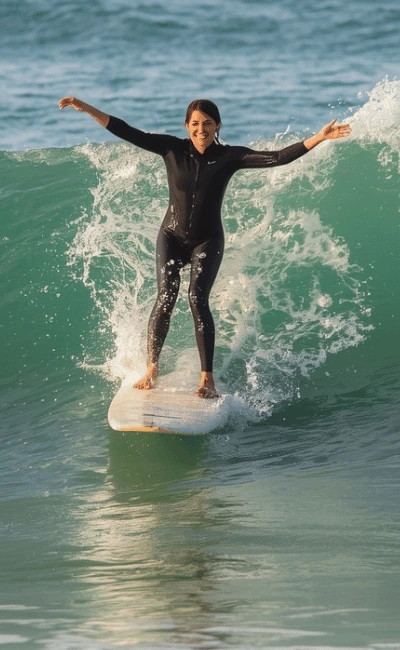 Woman in a black wetsuit riding a green wave on a white board with her arms spread wide